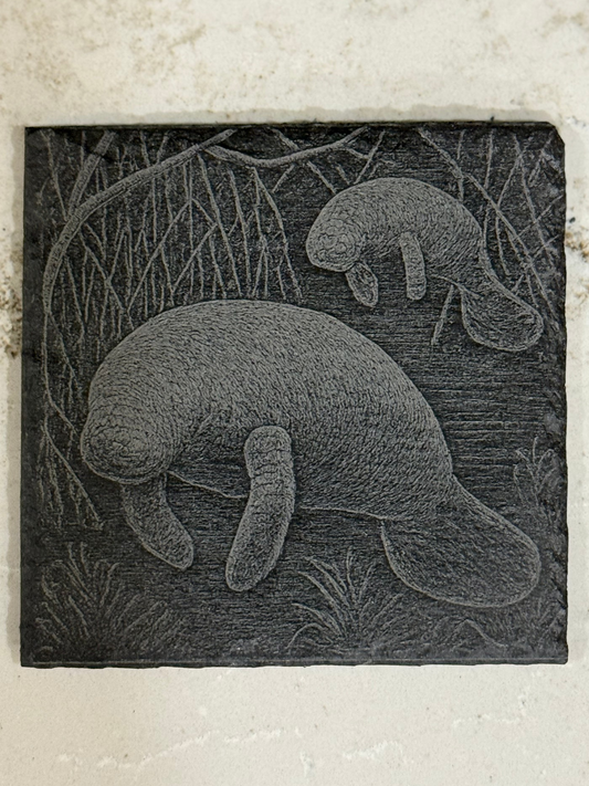 Carved stone plaque with two manatees in a natural setting on a textured surface.