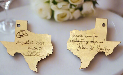 Two gold Texas-shaped charms with personalized text on a white surface with flowers in the background.